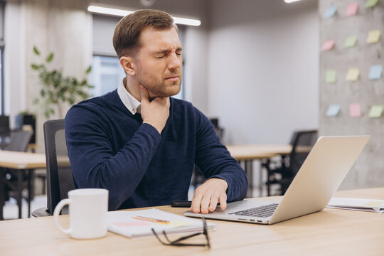 Office male worker grimacing, pressing neck while typing on laptop, suffering from throat pain or workplace related tension strain