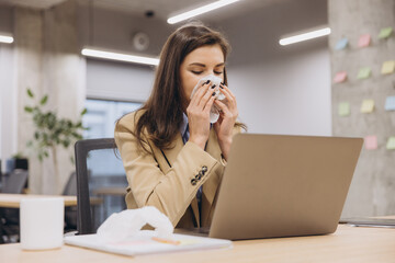 Obraz premium Professional businesswoman experiencing cold symptoms, wiping nose while working on laptop at clean, organized desk during office hours