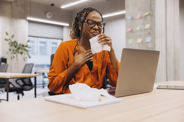 African American professional female covering mouth while sneezing near laptop, working in contemporary corporate workspace with sticky notes