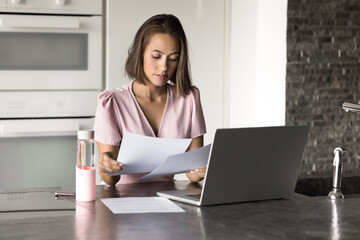 Young 25s woman sits at kitchen counter with laptop, focused on reviewing important documents,...