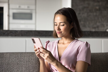 Young woman holding her smartphone seated on couch at home, responding to messages, learning new skills through platform, busy in content creation, editing videos or photos for blog. Modern tech usage