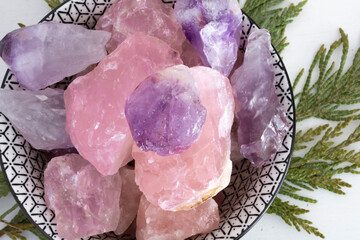 A top view image of a black and white bowl containing large, rough crystals of amethyst and rose quartz. The amethyst crystals are a rich purple, while the rose quartz crystals are a soft pink. 