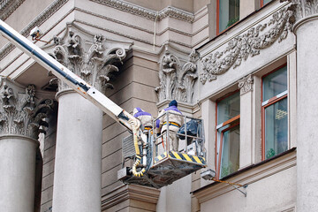 Workers on lift platform paint facade, facade restoration on historic building with ornate columns,...
