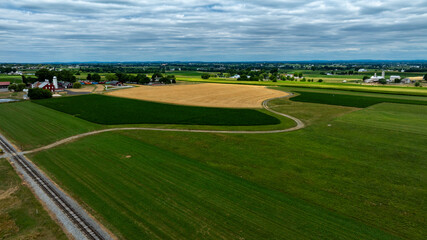 Aerial view captures extensive farmland with lush green fields and golden crops under a cloudy sky. Railroad tracks bisect the landscape, enhancing the serene rural setting.