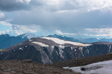 Dramatic vast view from sunlit high stony pass to big rocky top with snowfield, large snowy mountain range silhouette with pointy peaks far away under cloudy rainy sky. Sheer crags and peaked tops.