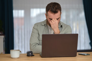 Tired man working overtime, sitting at the desk in front of a laptop and rubbing eyes with hands.