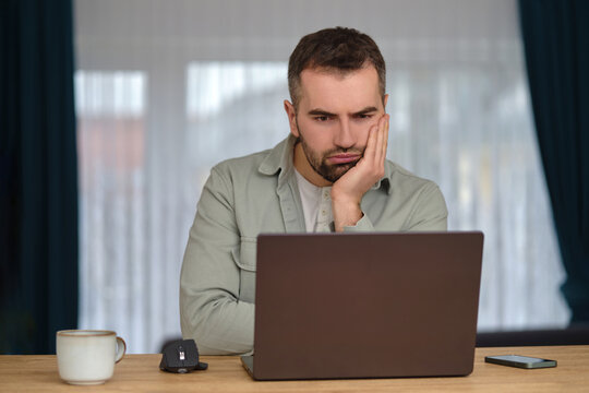 Bored man working overtime, sitting at the desk in front of a laptop and annoyed looking on the screen.