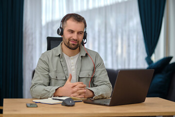 A happy man working at home office, wearing wireless headphones, having a video call with clients on a laptop, customer service or contact us support 