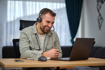 A happy man working at home office, wearing wireless headphones, having a video call with clients...