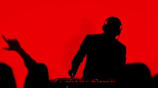 Silluette of a DJ playing music, red monotone background as the DJ switches knobs on the mixer and the crowd dancing