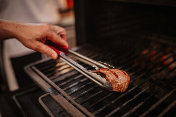 Close-up of a professional chef holding a charcoal-grilled lamb chop with tongs, with visible char marks