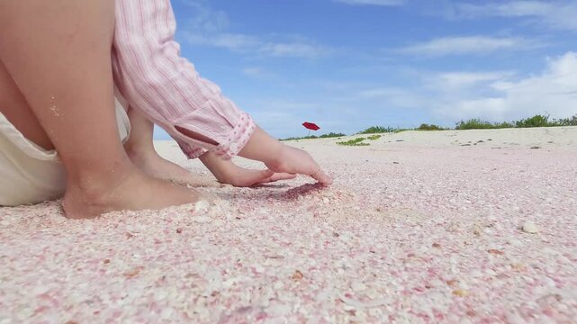 A young girl with red sunglasses and a striped shirt plays on the vibrant pink sand beach of Barbuda under a bright, clear sky. A red umbrella stands in the distance.