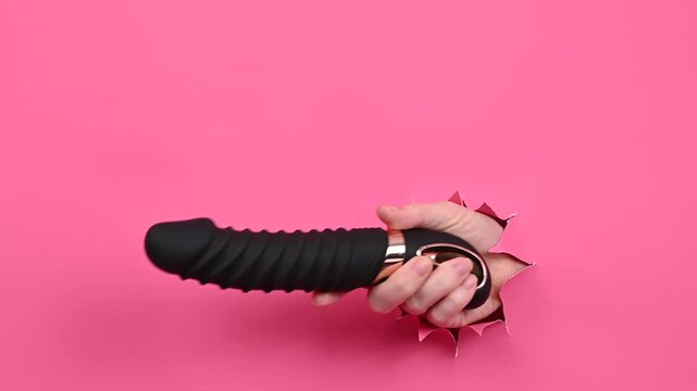 Female hand sticking out of pink paper background with black textured vibrator. Copy space. 