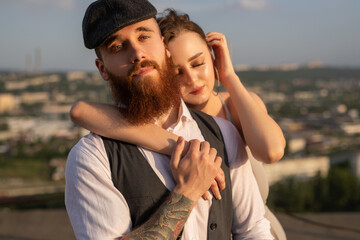 Fototapeta premium A young couple shares a romantic moment on a rooftop during sunset, embracing tenderly. The cityscape in the background adds depth, while the warm evening light highlights their affection and