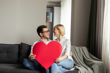 A joyful couple sit on a cozy couch, holding a large red heart symbol, celebrating love and...