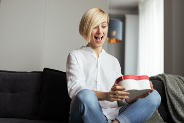 A cheerful woman sitting on a couch happily discovering a heartfelt gift during Valentine's...