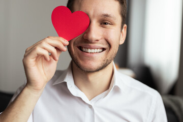 Cheerful man in a white shirt holds a red heart symbol, expressing joy and affection. Perfect...