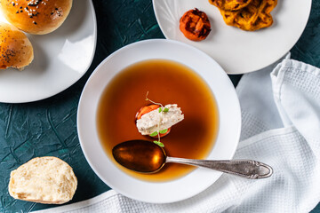 Soup or consomme, thick meat broth with carrots, chicken dumplings and microgreens, served in a white plate on a dark green background with sesame seed buns, napkin and spoon. View from above