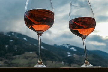 Two glasses of vibrant orange wine stand in sharp focus. Reflections shimmering against a blurred backdrop of the Austrian Alps in Matrei. Moment of relaxation and appreciation. Captured in East Tyrol