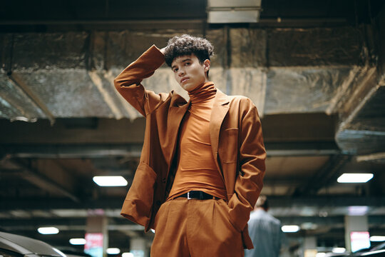 Stylish young man poses in an urban parking garage wearing an orange outfit with a brown blazer, showcasing modern fashion trends against a gritty backdrop