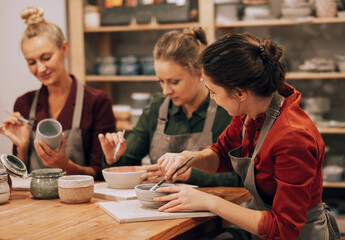 Three cheerful young women friends are painting ceramics in a pottery workshop. Have fun doing art.