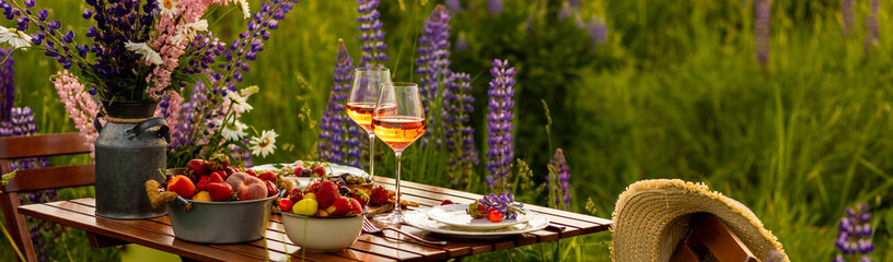 Banner. Two glasses of rose wine, flowers in a vase, silverware, fruits. Sunset, golden hour, atmospheric candlelight. Romantic table decor for a loving couple on blooming meadow with purple lupines