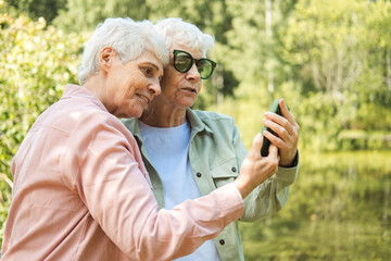 Two Caucasian pretty senior women smiling to smartphone camera while taking selfie photos near lake in the park.