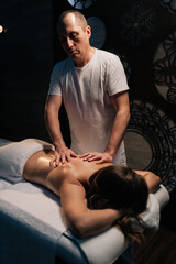 Vertical portrait of masseur giving relaxing back massage to young woman client lying down on massage table in a spa with warm candlelight creating peaceful atmosphere. Concept of luxury massage.