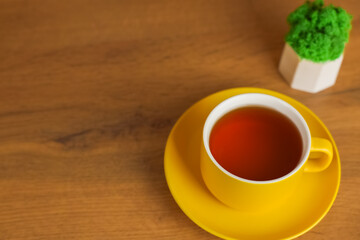 A top view of a yellow cup with a saucer in which black tea is poured stands on a wooden table on the left with space for text, next to it there is a plant in a white pot. Concept of tea drinking