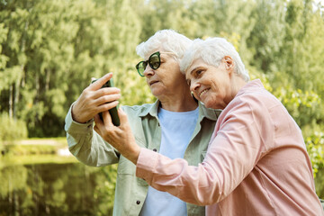 Two Caucasian pretty senior women smiling to smartphone camera while taking selfie photos near lake in the park.