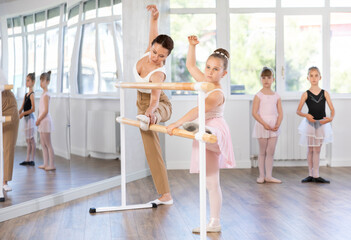 Fototapeta premium Adult woman teacher helps girl dancer stretch at ballet barre in studio