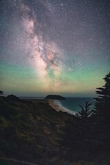 Big Sur Lighthouse over the sea with Milky Way in the sky. Serene landscape of mountains, trees, and water under a dark, starry sky