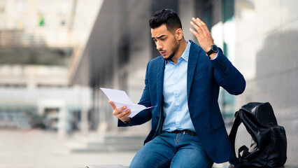 Emotional arabic guy young entrepreneur with backpack sitting on the street next to office...