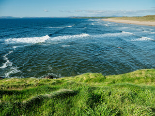 Green grass grows on cliff and amazing waves and sandy Bundoran beach in Ireland on warm sunny day. Popular tourist and surfers area. Blue cloudy sky. Irish landscape. Rich saturated color.