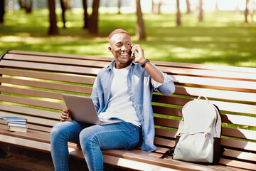 Fototapeta premium E-learning concept. Funky black student with laptop talking on smartphone on bench at park. African American guy talking to his college tutor while watching educational video on pc, outside