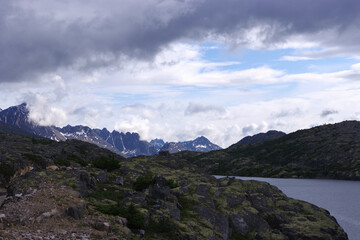 Sharp, jagged peaks rise into the cloud-covered sky, framing a serene alpine lake in the foreground. 