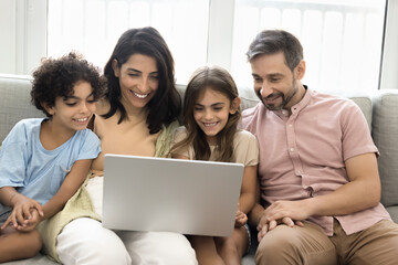 Beautiful family sitting on sofa using laptop, enjoying time together on internet, watching entertaining program, educational content, ordering goods, buy tickets, booking hotels, planning vacations