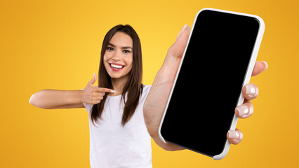 Try This App. Portrait of smiling lady holding smartphone with black blank screen in hand, showing device close up to camera and pointing finger at gadget with empty free space for mock up, banner