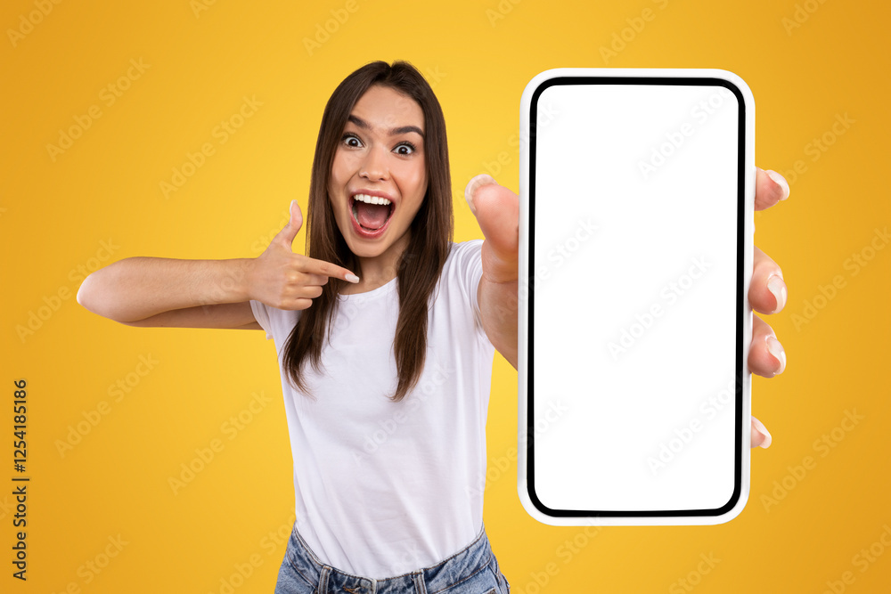 Wall mural Great Mobile Offer. Excited Lady Pointing Finger At Smartphone In Her Hand, Emotionally Reacting To New App, Overjoyed Millennial Woman Standing Isolated Over Orange Studio Background, Panorama