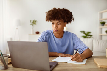 Studying from home concept. Serious black teen guy taking notes while participating in online lesson on laptop. Cool African American youth making homework, speaking to his tutor on web