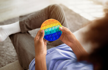 Cropped view of black teenager playing with antistress POP IT toy, sitting on sofa at home,...