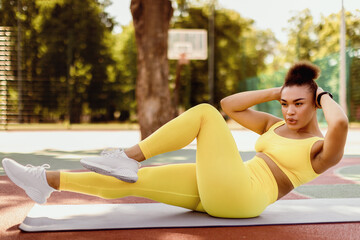 Obraz premium Weight Loss Concept. Confident Sportive Young Black Woman Doing Side Bicycle Crunches Bringing Elbow Toward Knee With Raised Leg On Yoga Mat, Training Outside At Court. Pilates And Warm Up Concept