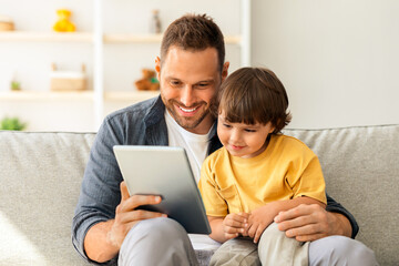 Fototapeta premium Child development content. Young positive father showing digital tablet to his cute little son, kid enjoying educational show online, sitting on sofa ta home