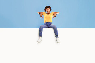 Excited young African American lady sitting on top of blank white paper banner, pointing down at copy space, advertising your service on blue studio background. Mockup for ad design