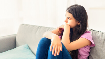 Stressed Indian teenage girl sitting on sofa at home, looking aside, feeling upset and lonely,...