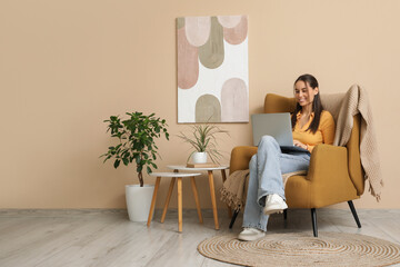 Young woman using laptop and sitting in armchair near beige wall in interior of living room