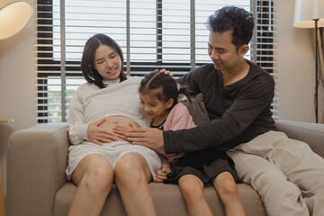 lovely family spend time together in their house, a pregnant mother sits on the couch with her husband and daughter enjoying a happy time together on a weekend