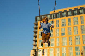 Fototapeta premium Kid swinging on chain swing on city kids playground. Swing ride. Cute child having fun on a swing on summer sky background. Kids playing in park. Blonde little boy swings at kid playground.
