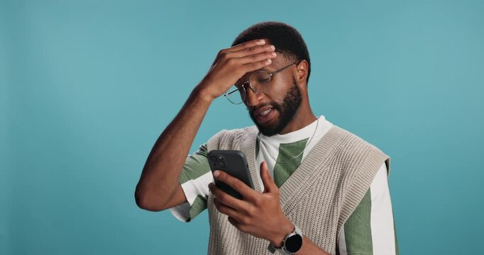 Frustrated, black man and stress with phone for bad news, debt or low credit score on a blue studio background. Male person, African user or model with migraine on mobile smartphone for bankruptcy