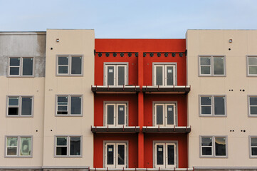 A modern apartment building under construction featuring a mix of beige and red exterior walls, multiple balconies, and large windows as part of an urban residential development proj